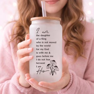 May include: Frosted glass tumbler with a bamboo lid and a clear straw. The tumbler features the text "I am the daughter of a King" with a floral illustration. The tumbler is held by a person wearing a pink sweater.