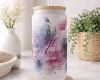 May include: A frosted glass tumbler with a bamboo lid and a clear straw, featuring a watercolor floral design in shades of pink and blue. The text "Be still and know that I am God" is printed in a dark pink script font across the center of the cup.