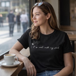 May include: Black t-shirt featuring the text "It is well with my soul" in white script, accompanied by a dandelion design. The shirt has a crew neck and short sleeves. A coffee cup rests on a wooden table near a window.