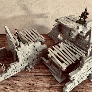 May include: A set of grey miniature stone ruins featuring crumbling walls and wooden plank roofs. Three small painted soldier figurines are positioned on the structures, with one standing on the roof and two others on the wall sections.