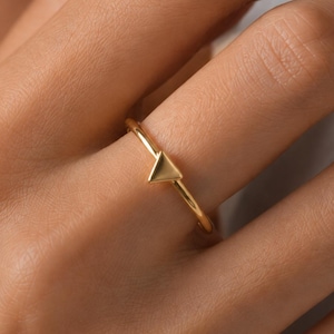 May include: A thin gold ring featuring a small, smooth, polished triangle charm centered on the band. The metal has a bright, reflective finish, and the geometric triangle element is integrated directly into the slender, circular ring band.