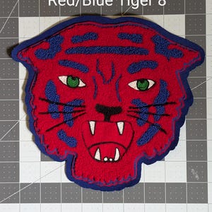 May include: A red and blue tiger patch with green eyes and white teeth. The patch is approximately 8 inches in size and features the text "Red/Blue Tiger 8"" in white. The tiger's face is detailed with blue accents.