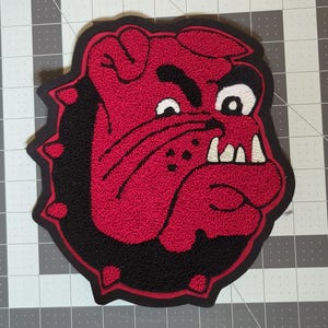 May include: A red and black embroidered patch featuring a bulldog's head. The bulldog has white teeth, a black eye, and a black eyebrow. The patch has a black border with red accents.