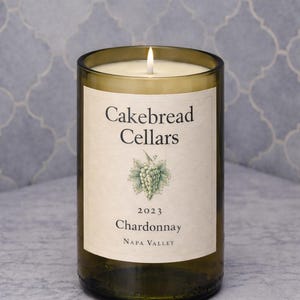 May include: A lit candle in a repurposed wine bottle. The label reads "Cakebread Cellars 2023 Chardonnay Napa Valley" with a grape cluster illustration. The candle is in a clear glass bottle with a light-colored wax.