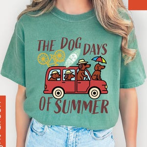 May include: Light green t-shirt with a graphic of dachshunds in a red car, a bicycle, and the text "THE DOG DAYS OF SUMMER". The shirt is made of a soft material and is suitable for casual wear.