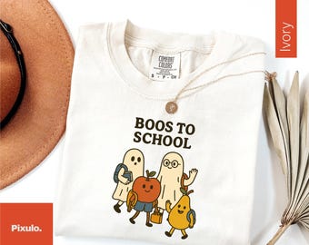 May include: Ivory-colored t-shirt with the text "Boos to School" and a graphic of cartoon ghosts, an apple, and a pear, all wearing backpacks. A gold necklace is draped across the shirt. A brown hat and dried leaves are in the background.