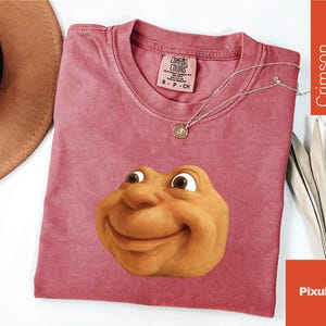 May include: Crimson-colored t-shirt with a smiling cartoon face graphic. The shirt has a crew neck and short sleeves. A silver necklace with a pendant is on the shirt. The shirt is labeled "Comfort Colors" with size information.