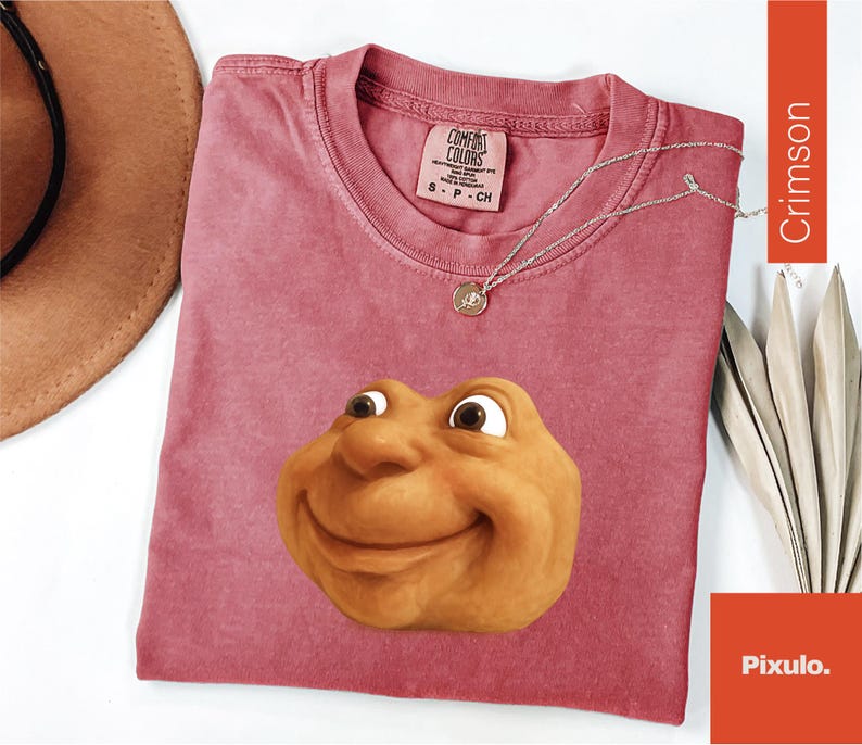 May include: Crimson-colored t-shirt with a smiling cartoon face graphic. The shirt has a crew neck and short sleeves. A silver necklace with a pendant is on the shirt. The shirt is labeled "Comfort Colors" with size information.