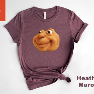 May include: Heather maroon t-shirt featuring a smiling, cartoonish face with brown skin and large eyes. The shirt is knotted at the bottom. The words "Heather Maroon" are printed in a vertical format on the right side.