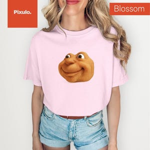 May include: A light pink t-shirt featuring a smiling, cartoonish face. The shirt has short sleeves and is paired with denim shorts. The word "Blossom" is printed in white on a red rectangle in the upper right corner.
