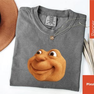 May include: A gray Comfort Colors t-shirt featuring a smiling, cartoonish face in shades of brown and tan. A silver necklace with a pendant rests on the shirt. The shirt is size S-P-CH.