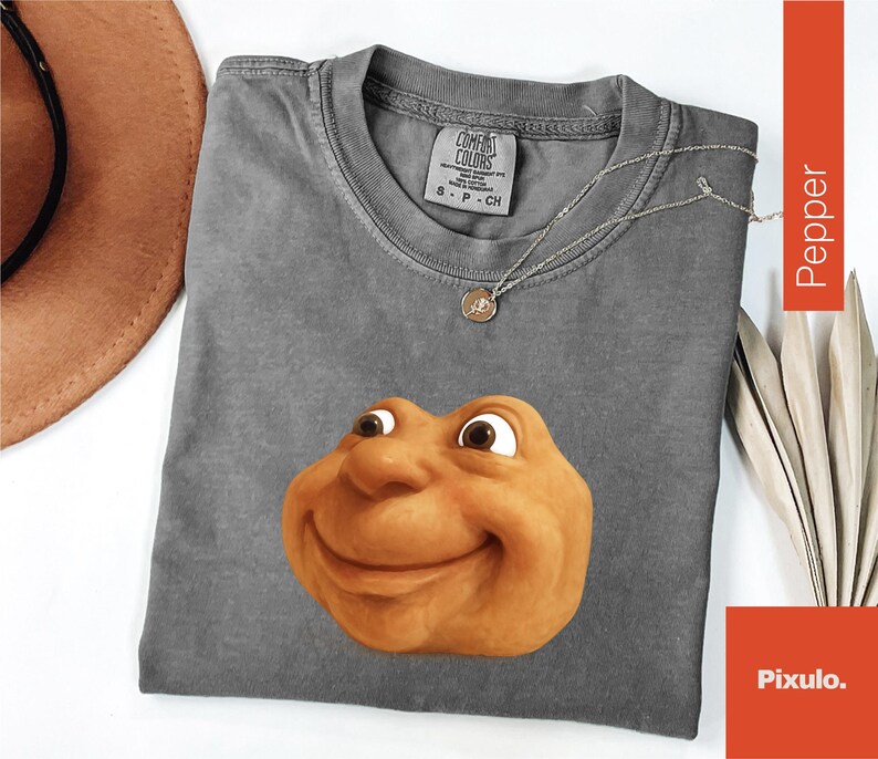 May include: A gray Comfort Colors t-shirt featuring a smiling, cartoonish face in shades of brown and tan. A silver necklace with a pendant rests on the shirt. The shirt is size S-P-CH.