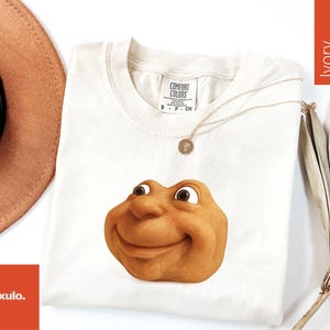 May include: Ivory-colored t-shirt with a smiling, cartoonish face graphic. The shirt has a crew neck and short sleeves. A gold necklace with a pendant is draped across the shirt. A brown hat and dried leaves are in the background.