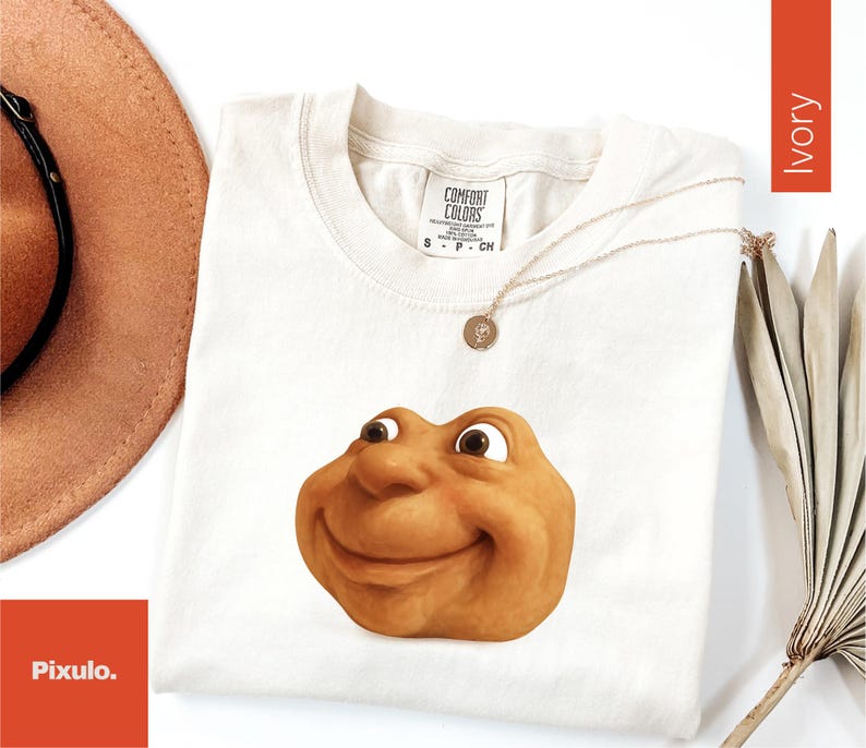 May include: Ivory-colored t-shirt with a smiling, cartoonish face graphic. The shirt has a crew neck and short sleeves. A gold necklace with a pendant is draped across the shirt. A brown hat and dried leaves are in the background.