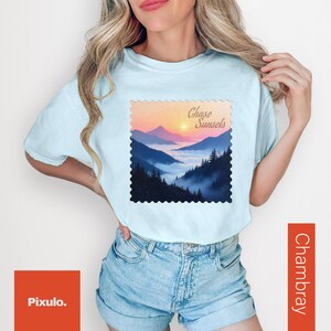 May include: Light blue t-shirt with a landscape graphic featuring mountains, a sunset, and the text "Chase Sunsets." The shirt is paired with denim shorts. The graphic is framed with a decorative border.