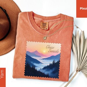 May include: A coral-colored t-shirt with a landscape graphic featuring mountains, trees, and a sunset, with the text "Chase Sunsets." A gold necklace and a brown hat are also visible.