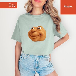 May include: A light green t-shirt featuring a smiling, cartoonish face with brown eyes and a tan complexion. The shirt has short sleeves and a relaxed fit. The word "Bay" is in the top left corner and "Pixulo." is in the top right corner.