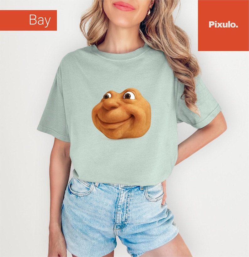 May include: A light green t-shirt featuring a smiling, cartoonish face with brown eyes and a tan complexion. The shirt has short sleeves and a relaxed fit. The word "Bay" is in the top left corner and "Pixulo." is in the top right corner.