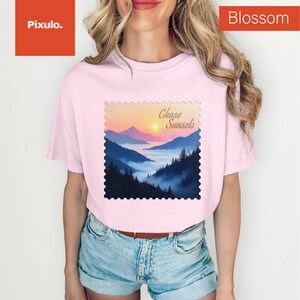 May include: A light pink t-shirt featuring a landscape design with the text "Chase Sunsets." The design depicts a sunset over mountains with a blue and orange color scheme. The shirt is a comfortable, casual style.