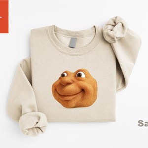 May include: Sand-colored sweatshirt featuring a smiling, cartoonish face. The face is a warm, earthy tone with large eyes and a friendly expression. The word "Sand" is printed in a dark color on the bottom right.