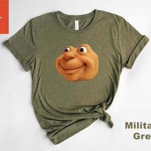 May include: Olive green t-shirt featuring a smiling, cartoonish face in shades of brown and tan. The shirt has short sleeves and a crew neck. The words "Military Green" are printed on the bottom right.