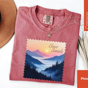 May include: Crimson-colored t-shirt with a crew neck and a graphic of a mountain landscape with the text "Chase Sunsets." A silver necklace with a pendant is on the shirt. The shirt is made by Comfort Colors.