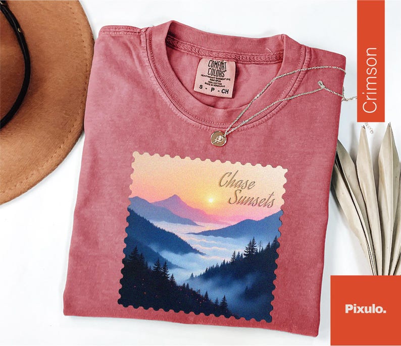 May include: Crimson-colored t-shirt with a crew neck and a graphic of a mountain landscape with the text "Chase Sunsets." A silver necklace with a pendant is on the shirt. The shirt is made by Comfort Colors.