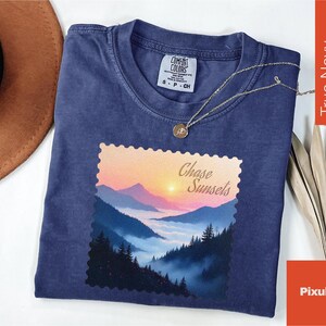 May include: A navy blue t-shirt with a landscape graphic featuring a sunset over mountains and the text "Chase Sunsets." The shirt has a crew neck and short sleeves. A gold necklace with a pendant is draped over the shirt.