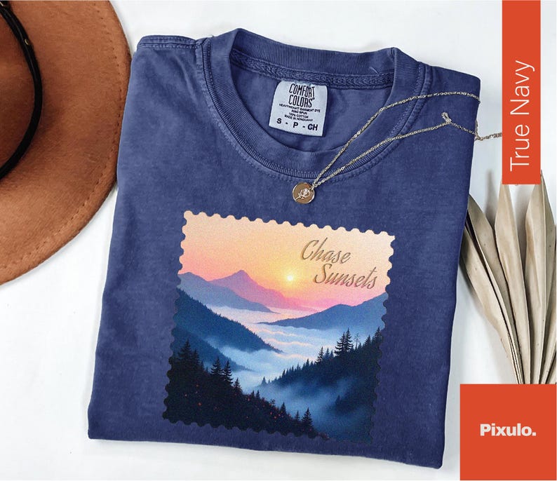May include: A navy blue t-shirt with a landscape graphic featuring a sunset over mountains and the text "Chase Sunsets." The shirt has a crew neck and short sleeves. A gold necklace with a pendant is draped over the shirt.
