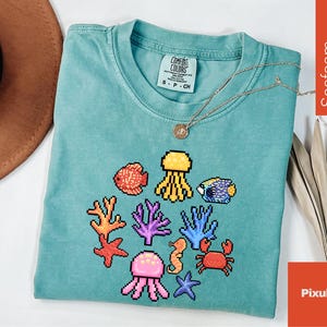 May include: Seafoam green t-shirt with a pixelated aquatic design featuring fish, jellyfish, coral, a seahorse, crab, and starfish. The shirt has a Comfort Colors tag and a gold necklace. The word "Seafoam" is printed vertically on the right side.