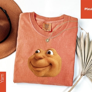 May include: A coral-colored t-shirt featuring a smiling, cartoonish face. The shirt has a crew neck and a tag that reads "Comfort Colors". A gold necklace with a pendant and a brown hat are also visible.