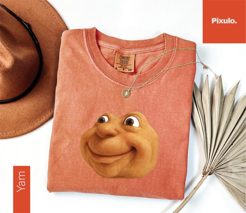 May include: A coral-colored t-shirt featuring a smiling, cartoonish face. The shirt has a crew neck and a tag that reads "Comfort Colors". A gold necklace with a pendant and a brown hat are also visible.