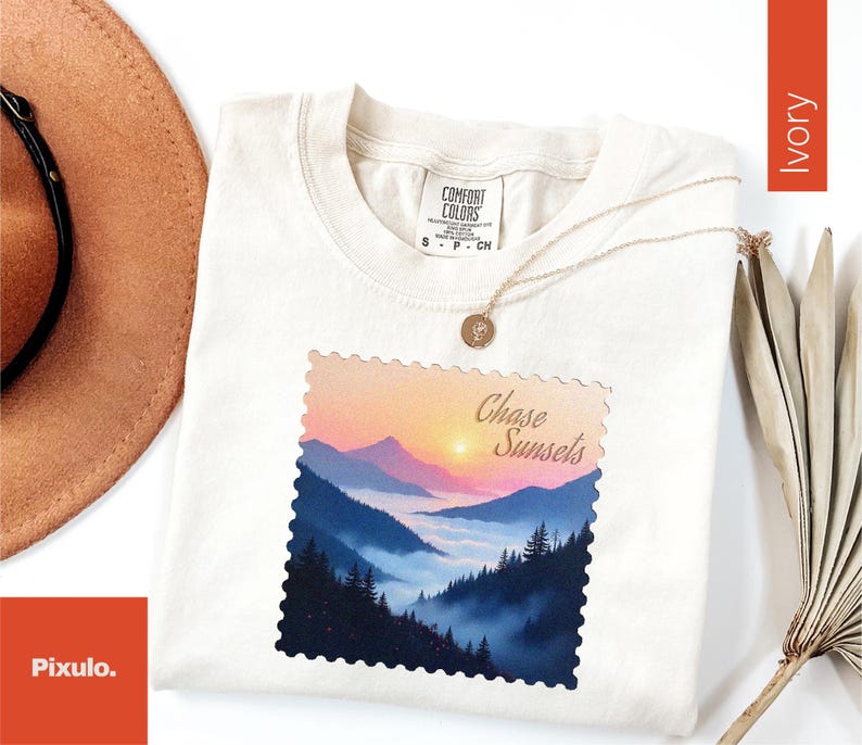 May include: Ivory-colored t-shirt with a mountain landscape graphic. The graphic features a sunset over a mountain range with the words "Chase Sunsets". A gold necklace with a pendant is draped over the shirt.