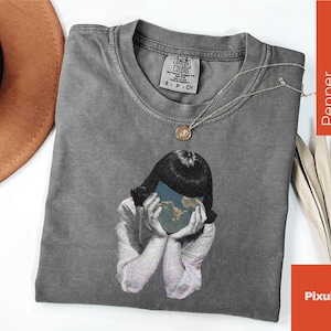 May include: A gray Comfort Colors t-shirt featuring a graphic of a person holding a globe in front of their face. A gold necklace with a pendant rests on the shirt. A brown hat is visible in the upper left corner.