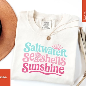 May include: Ivory-colored t-shirt with the words "Saltwater, Seashells, Sunshine" in turquoise and pink. A gold necklace with a pendant rests on the shirt. A brown hat and dried palm leaves are in the background.