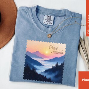 May include: Blue jean colored t-shirt with a landscape graphic featuring mountains and a sunset. The graphic is in a postage stamp style and reads "Chase Sunsets". A silver necklace with a pendant is draped across the shirt.