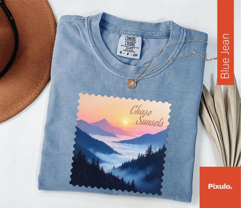 May include: Blue jean colored t-shirt with a landscape graphic featuring mountains and a sunset. The graphic is in a postage stamp style and reads "Chase Sunsets". A silver necklace with a pendant is draped across the shirt.