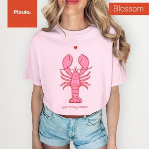 May include: A light pink t-shirt featuring a pink lobster illustration with the text "you're my lobster" underneath. A small red heart is above the lobster. The word "Blossom" is in a red box in the upper right corner.