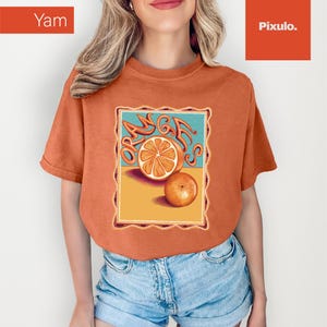 May include: Orange-colored t-shirt featuring a graphic of an orange slice and a whole orange with the word "ORANGE" in stylized lettering. The design is framed within a rectangular border. The model is wearing blue denim shorts.