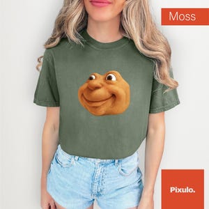 May include: A moss green t-shirt featuring a cartoonish, smiling face. The graphic is a warm, golden brown color. The shirt is paired with blue denim shorts. The word "Moss" is in a red box in the upper right corner.