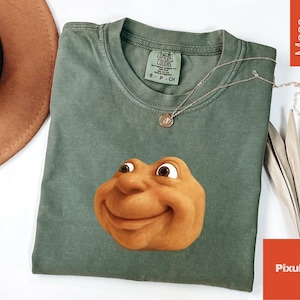 May include: A moss green t-shirt with a smiling cartoon face graphic. The shirt has a crew neck and short sleeves. A silver necklace with a pendant is draped on the shirt. The shirt has a Comfort Colors tag.