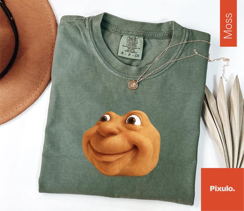 May include: A moss green t-shirt with a smiling cartoon face graphic. The shirt has a crew neck and short sleeves. A silver necklace with a pendant is draped on the shirt. The shirt has a Comfort Colors tag.
