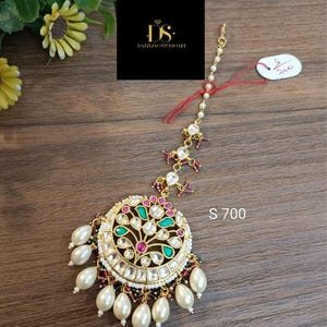 Puede incluir: Un tocado dorado adornado con un colgante circular con piedras preciosas verdes, rosas y blancas. El colgante está adornado con perlas de imitación y cuelga de una delicada cadena. La etiqueta dice "S 700".