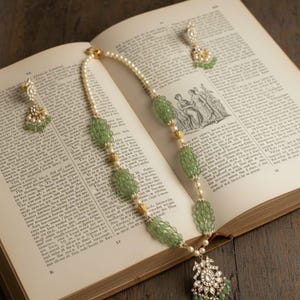 Op de afbeelding: Een ketting en bijpassende oorbellen set met groene kralen, parels en gouden accenten, gepresenteerd op een open boek. De ketting heeft een hanger met heldere stenen en groene accenten. De oorbellen hebben een vergelijkbaar ontwerp.