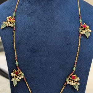 May include: Gold-toned necklace and matching earrings set. The necklace features a delicate chain with ruby and emerald accents, and clusters of pearls and gemstones. The earrings mirror the necklace design. The text "FW75" is visible.
