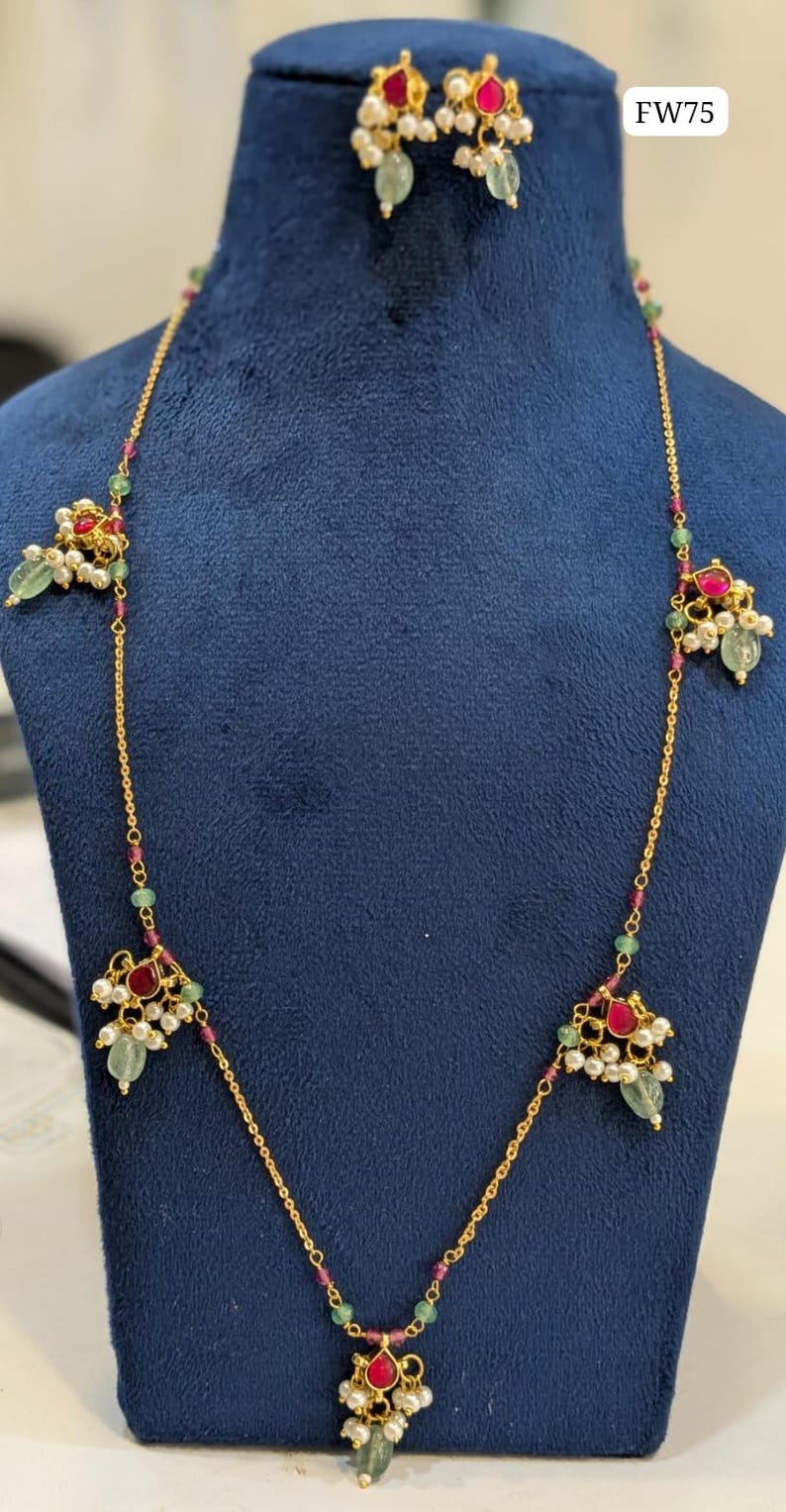 May include: Gold-toned necklace and matching earrings set. The necklace features a delicate chain with ruby and emerald accents, and clusters of pearls and gemstones. The earrings mirror the necklace design. The text "FW75" is visible.