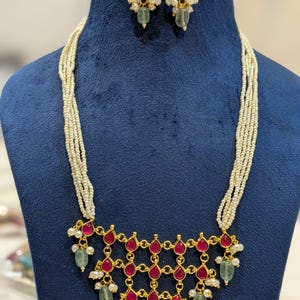 May include: A gold-toned necklace and earring set. The necklace features a geometric design with red teardrop-shaped stones, green stones, and small white pearls. The earrings mirror the design with red stones, pearls, and green stones. The necklace has multiple strands of small white beads.