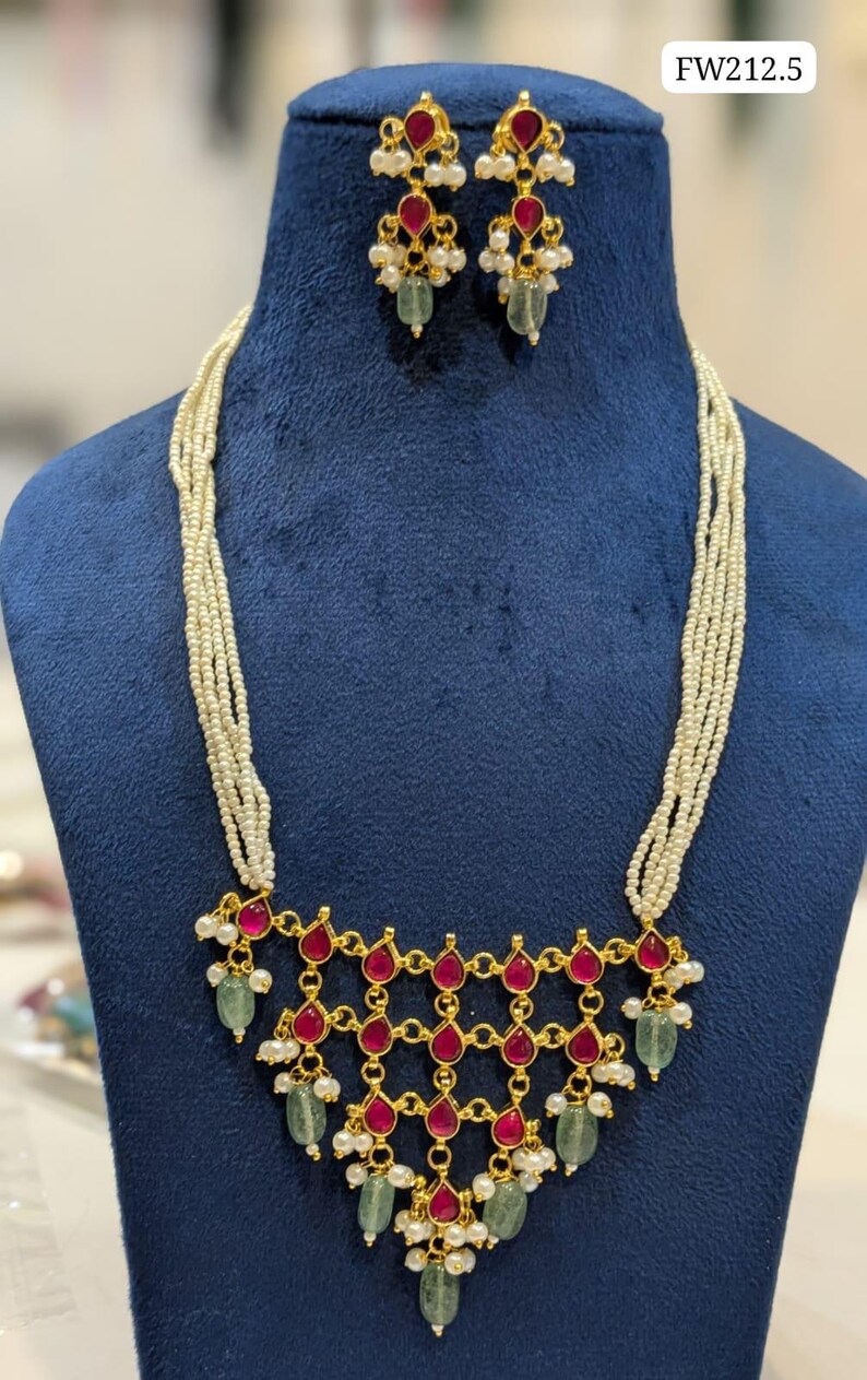 May include: A gold-toned necklace and earring set. The necklace features a geometric design with red teardrop-shaped stones, green stones, and small white pearls. The earrings mirror the design with red stones, pearls, and green stones. The necklace has multiple strands of small white beads.
