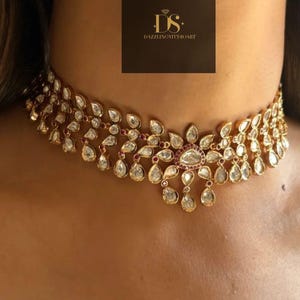 May include: A gold-toned choker necklace with clear and red gemstones. The necklace features a floral design with teardrop-shaped gems and a central cluster of stones. The necklace is worn around the neck.