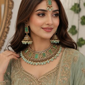 May include: A jewelry set featuring a gold-toned necklace, matching jhumka earrings, and a maang tikka. Each piece is adorned with rectangular mint green stones, small clear crystals, and dangling mint green beads set in an intricate metal frame.